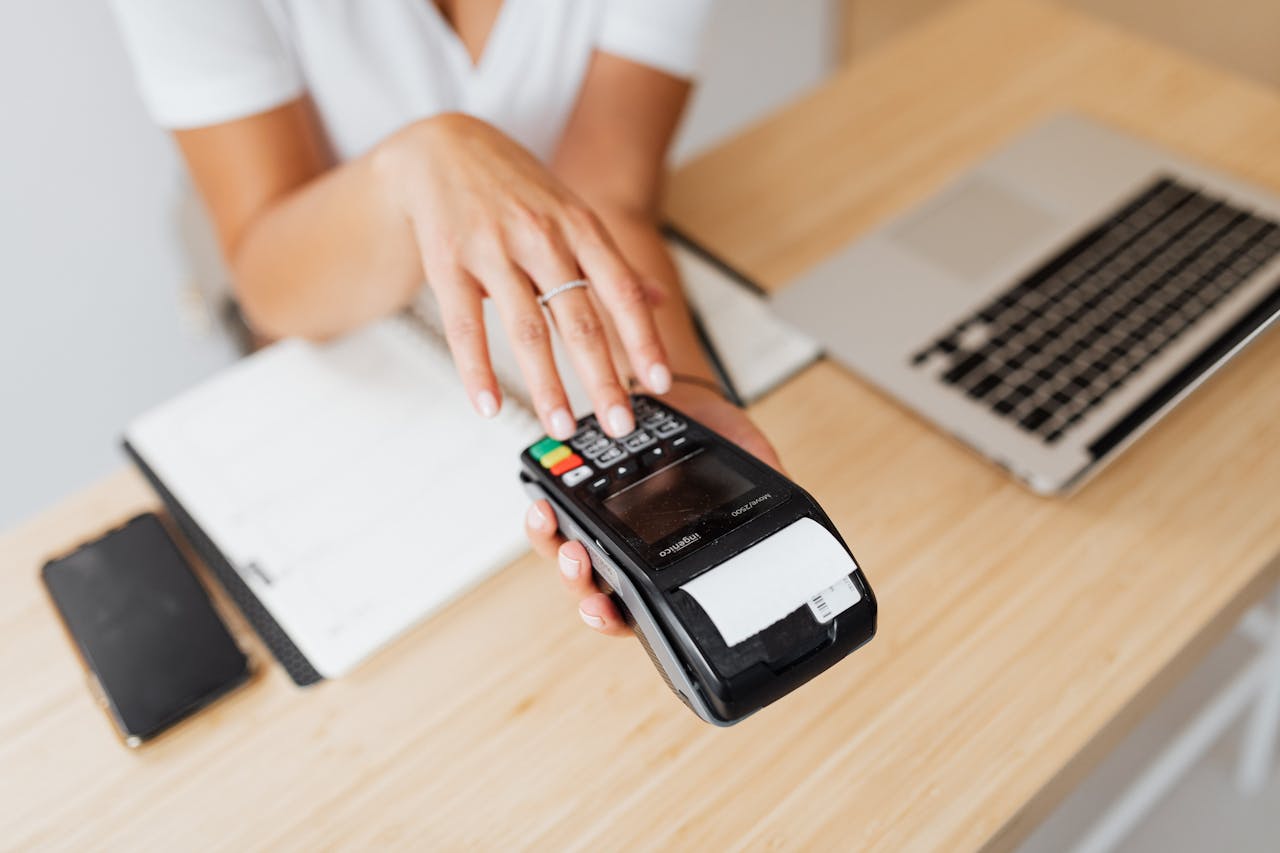 Close-up of a hand using a payment terminal beside a laptop on a wooden countertop.